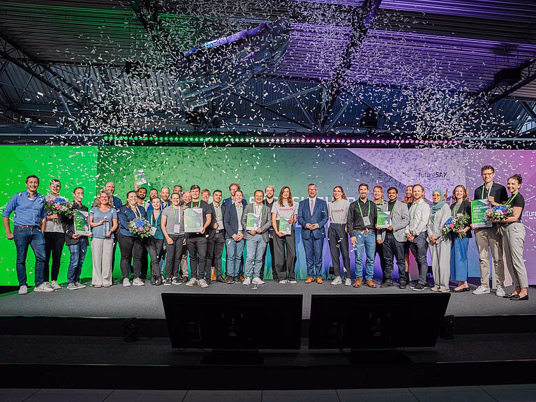 Eine gro&szlig;e B&uuml;hne bei der S&auml;chsischen Innovationskonferenz 2024. Auf der B&uuml;hne stehen mehrere Personen, die in einem feierlichen Rahmen ausgezeichnet werden. Im Hintergrund ist ein gro&szlig;es Banner mit dem Logo der Konferenz und der Aufschrift 'Innovationskonferenz 2024' zu sehen. Das Publikum im Vordergrund applaudiert den Preistr&auml;gern